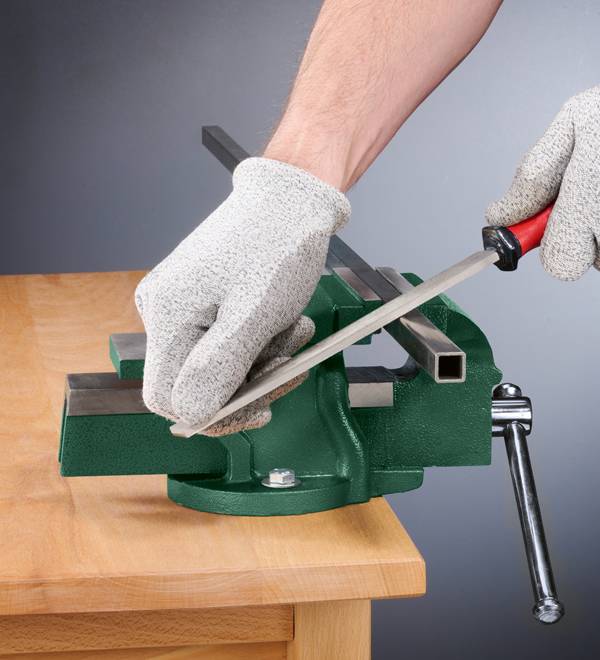 A person wearing gloves uses a file to smooth a metal bar held in a green vise.