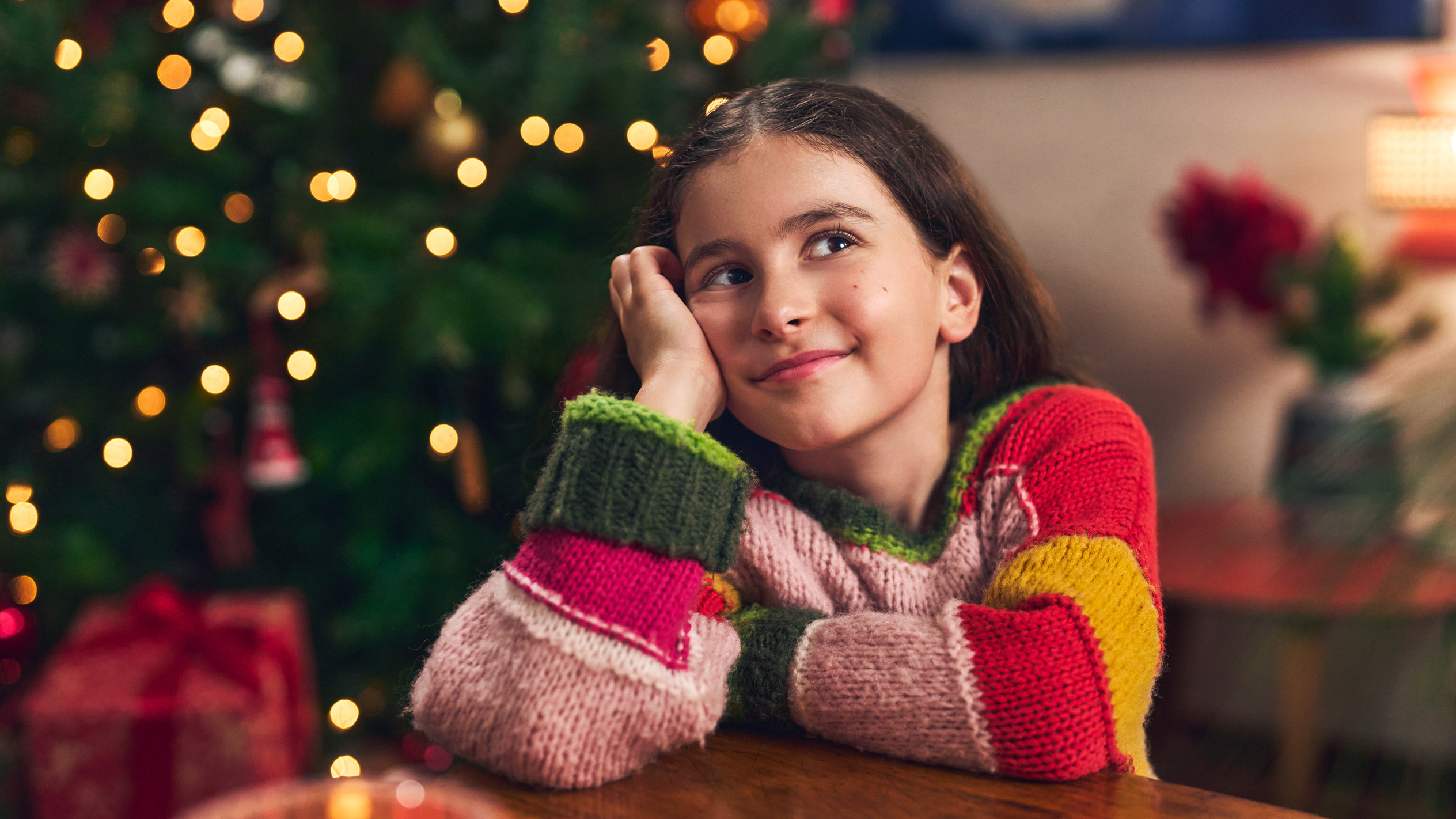Smiling girl in a colorful knitted sweater, dreaming by a Christmas tree.