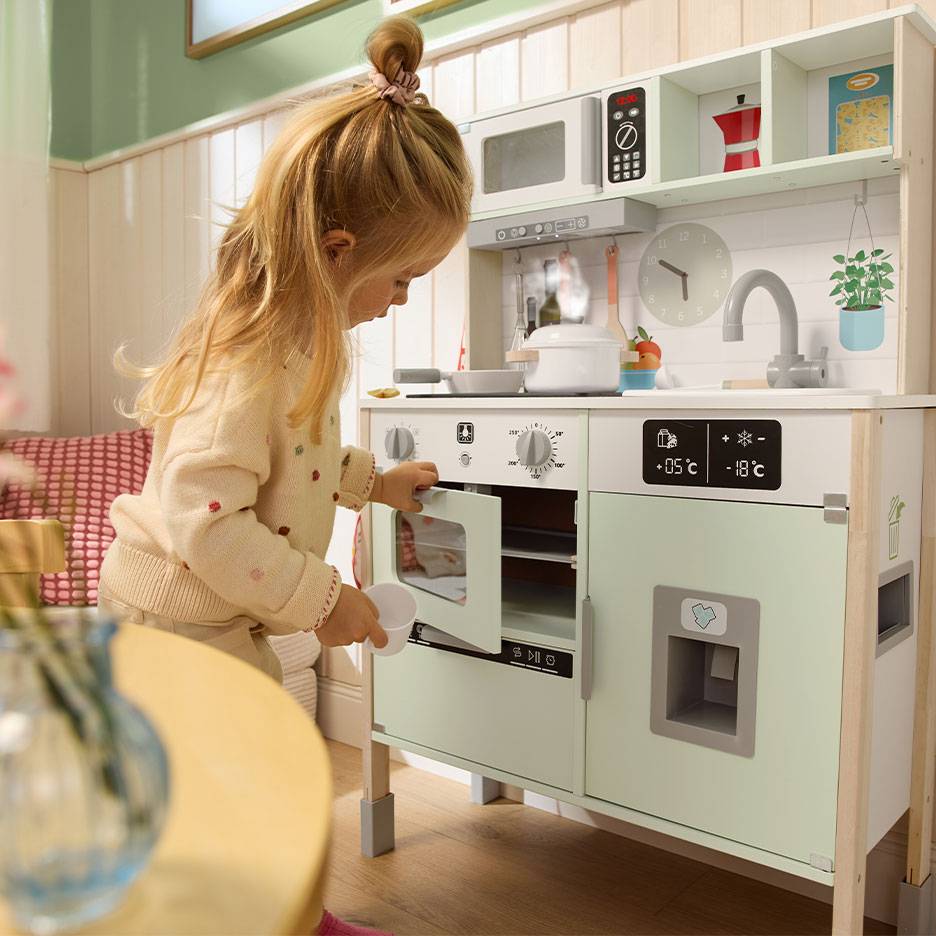 Child playing with a wooden toy kitchen, featuring an oven, microwave, and fridge.
