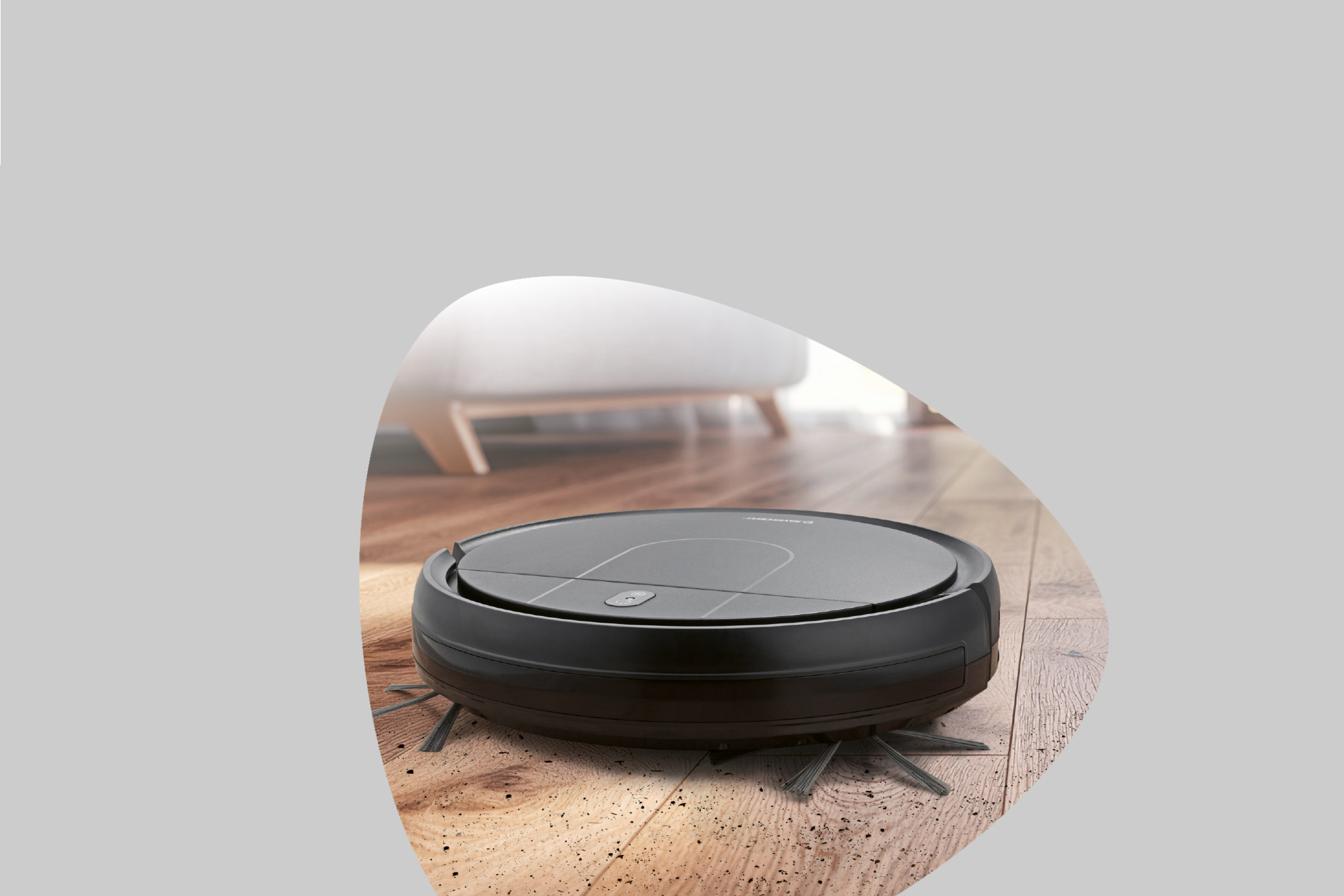 A robotic vacuum cleaner on a wooden floor, surrounded by dirt, with a blurred sofa in the background.