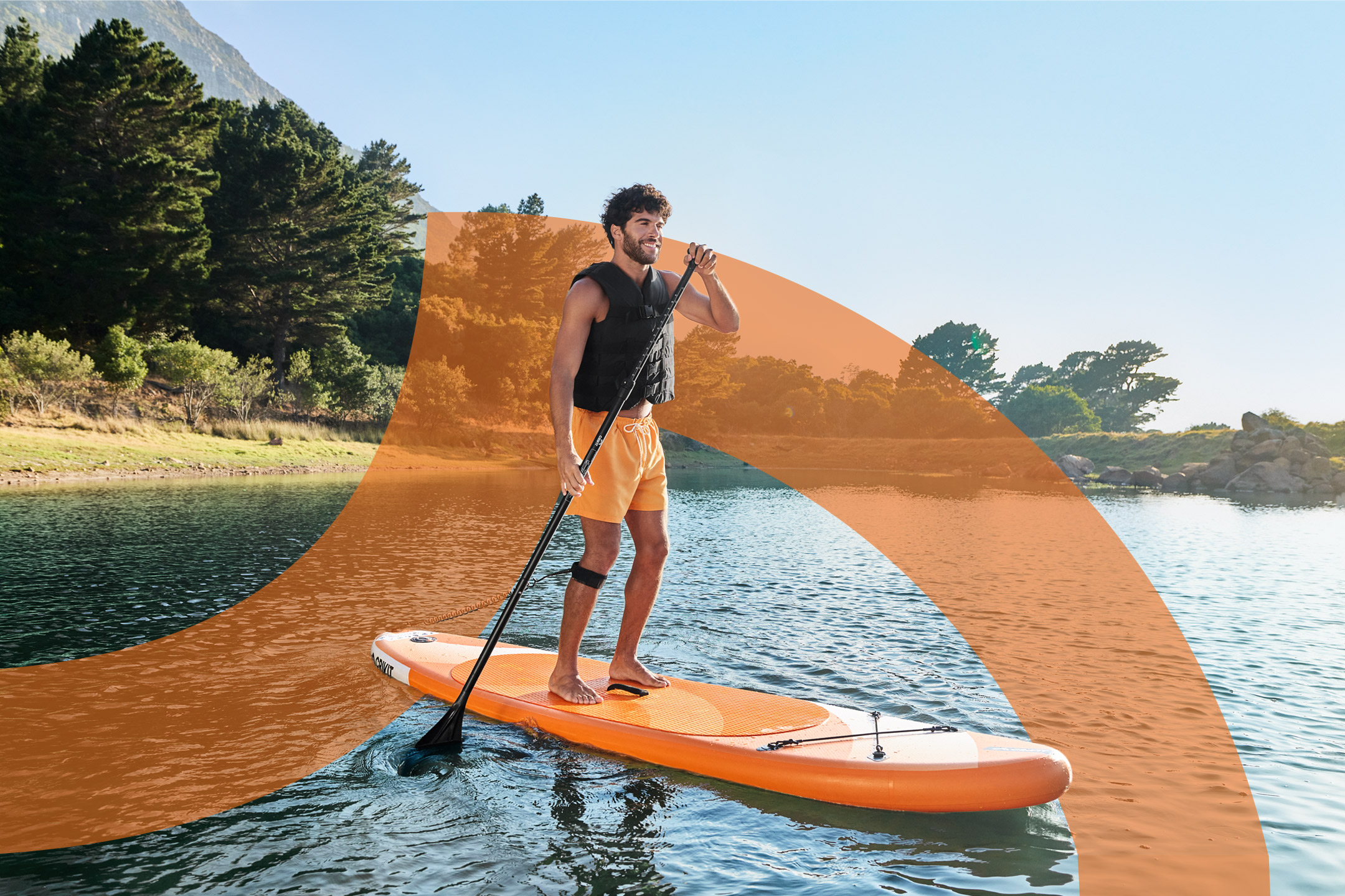 Man paddleboarding on a lake, wearing a life vest and orange shorts.