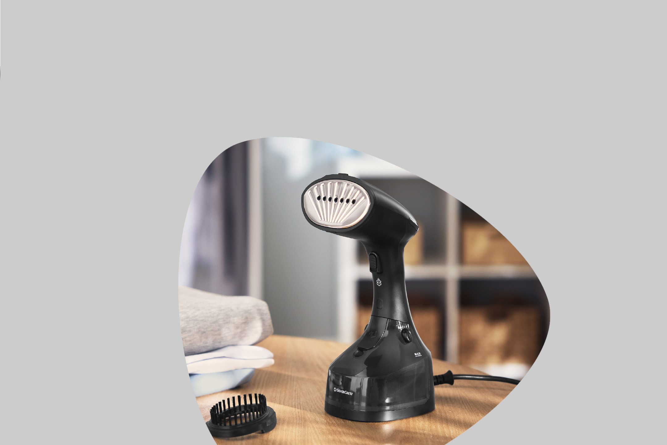 Black handheld garment steamer with a brush attachment on a wooden table next to folded clothes.