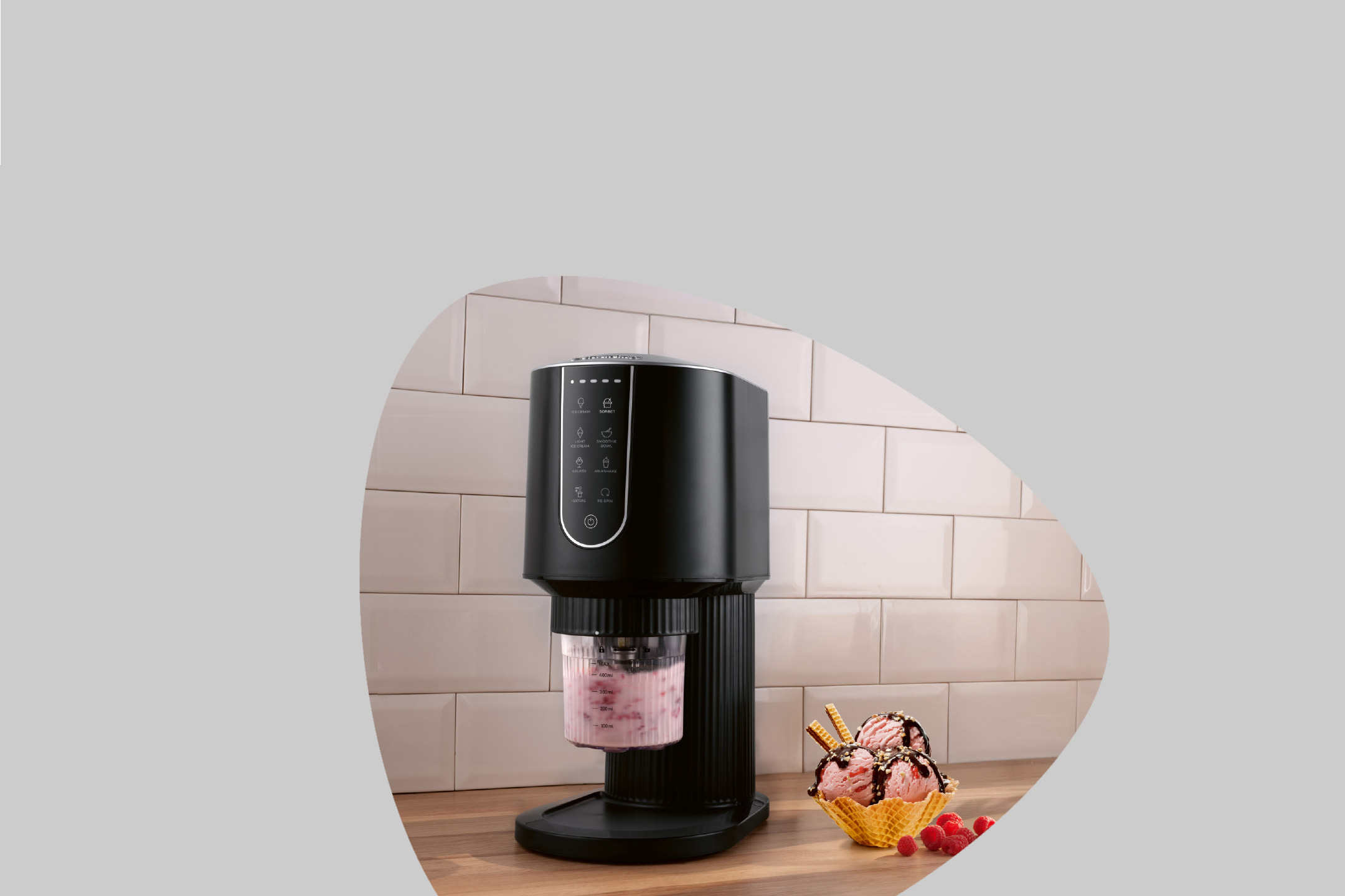 Black ice cream maker with strawberry ice cream in a cup, next to a waffle cone with ice cream.
