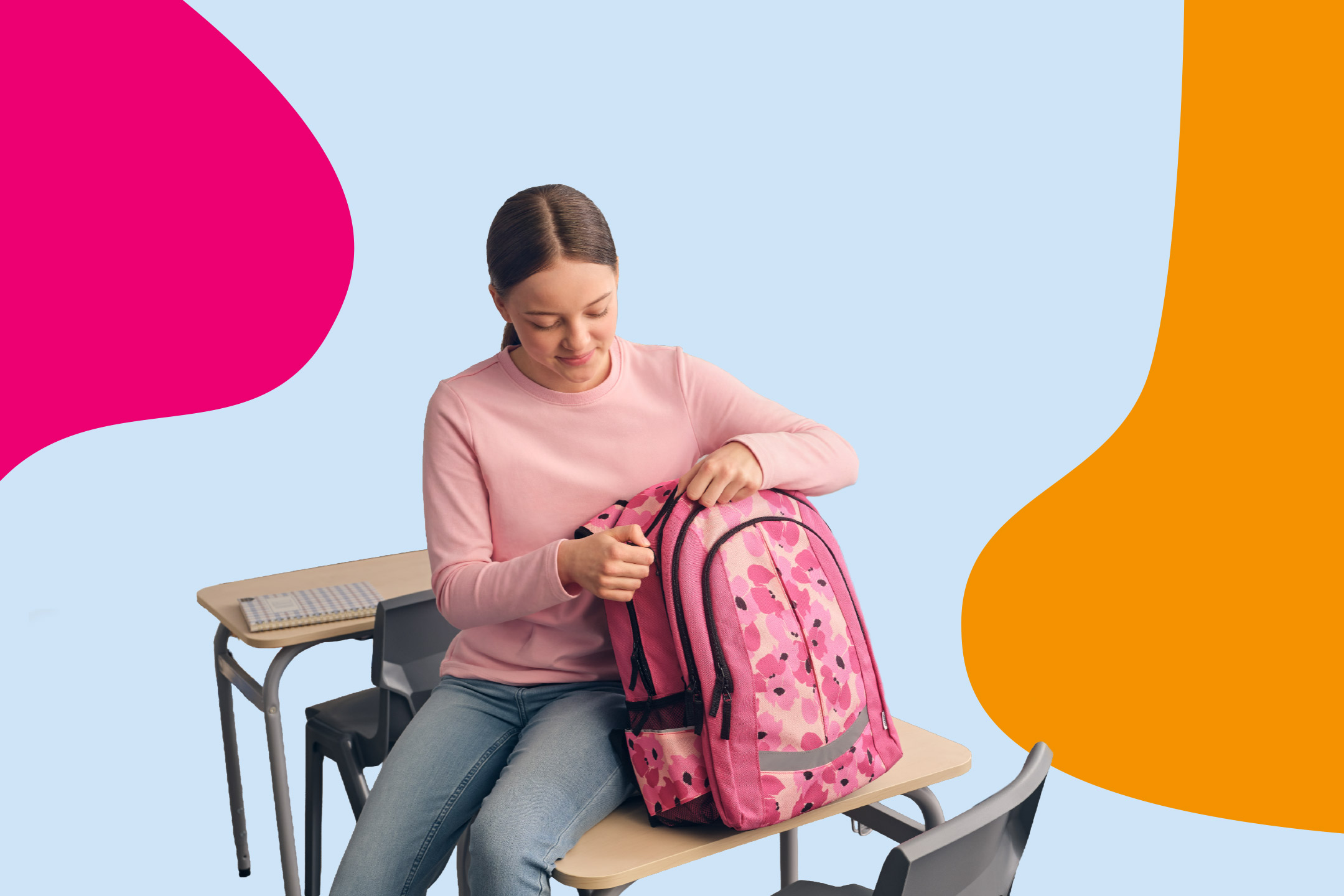 Girl in a pink sweatshirt and jeans, sitting at a school desk, opening a pink floral backpack.