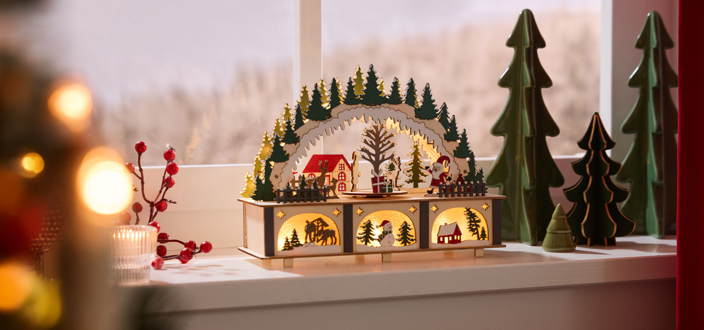 Christmas decorations: illuminated wooden landscape with houses and trees, red berries and candle.