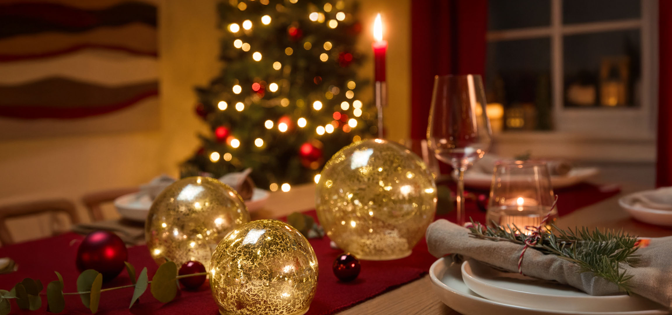 Christmas table setting with golden light globes, red baubles, and fir sprigs on a red tablecloth.