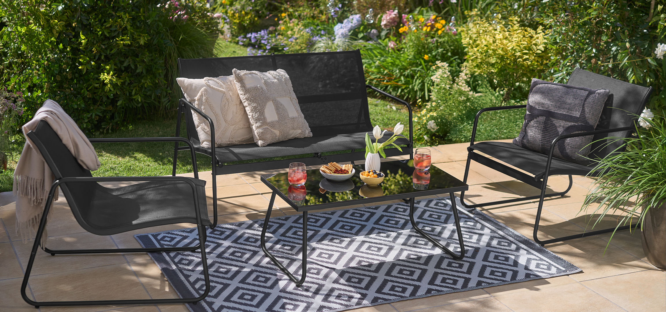 Outdoor furniture set with black chairs, bench, table, and rug in a garden.