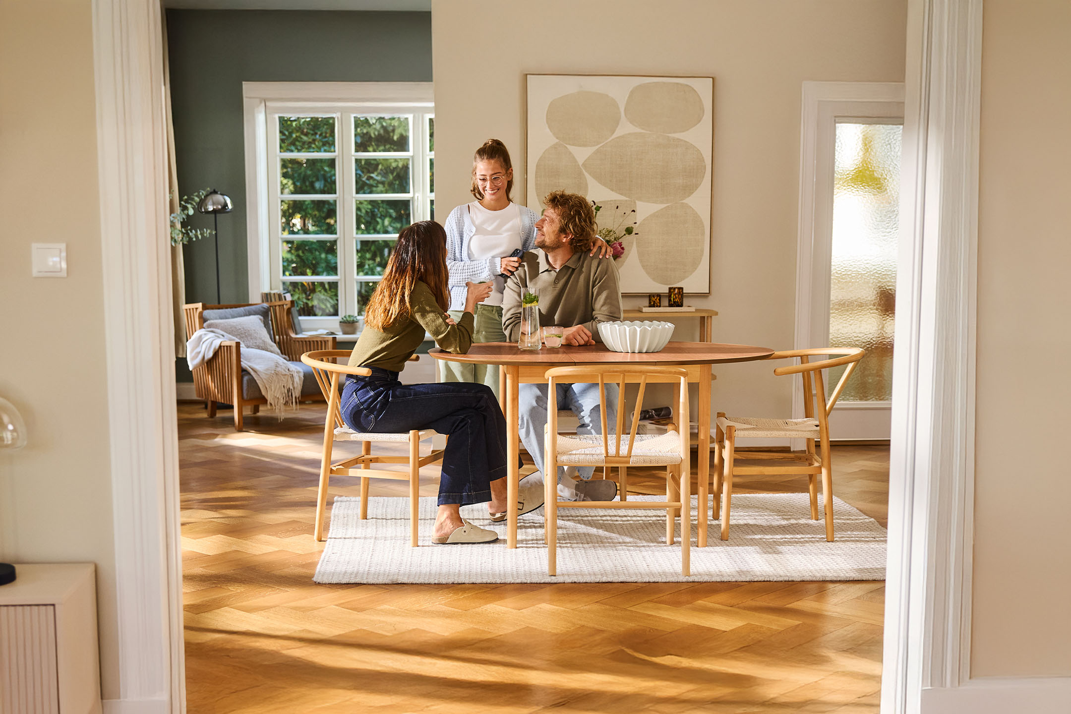 Three people gathered around a wooden dining table with chairs in a bright living space.