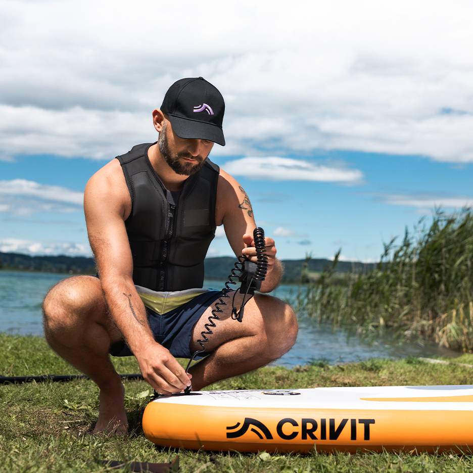 Man attaches safety leash to CRIVIT SUP board, wearing a life vest and cap.
