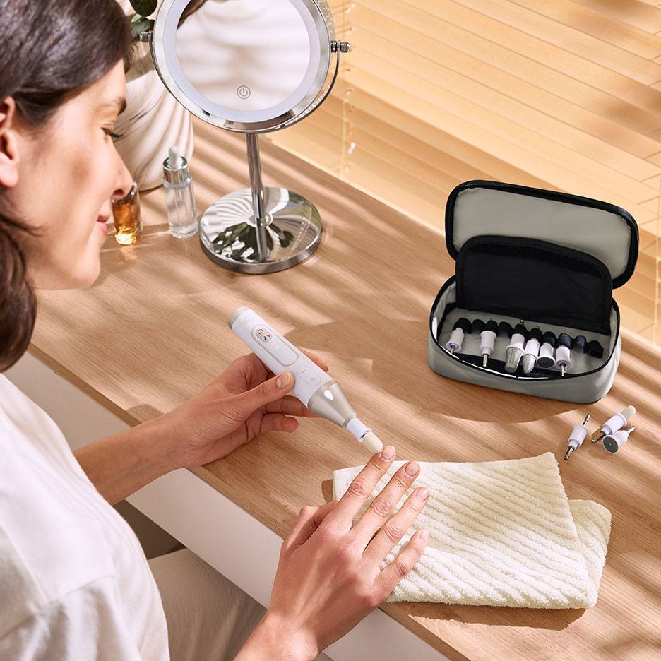 Woman using an electric nail file with attachments, next to a mirror and a case with accessories.
