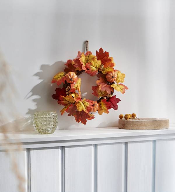 A fall-themed wreath with colorful leaves and small pumpkins hangs above a white mantelpiece.