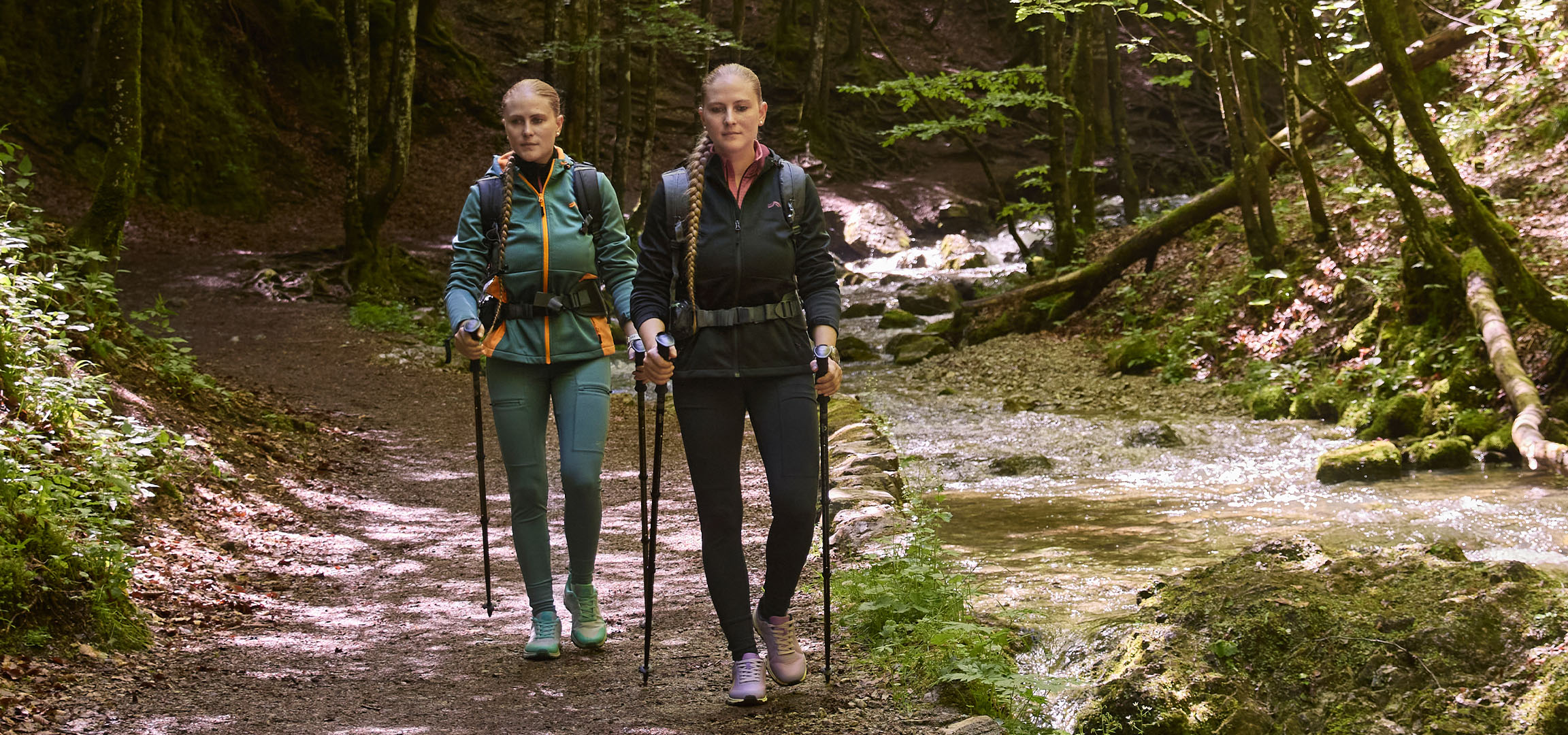 Two women hike along a stream in a forest using trekking poles.