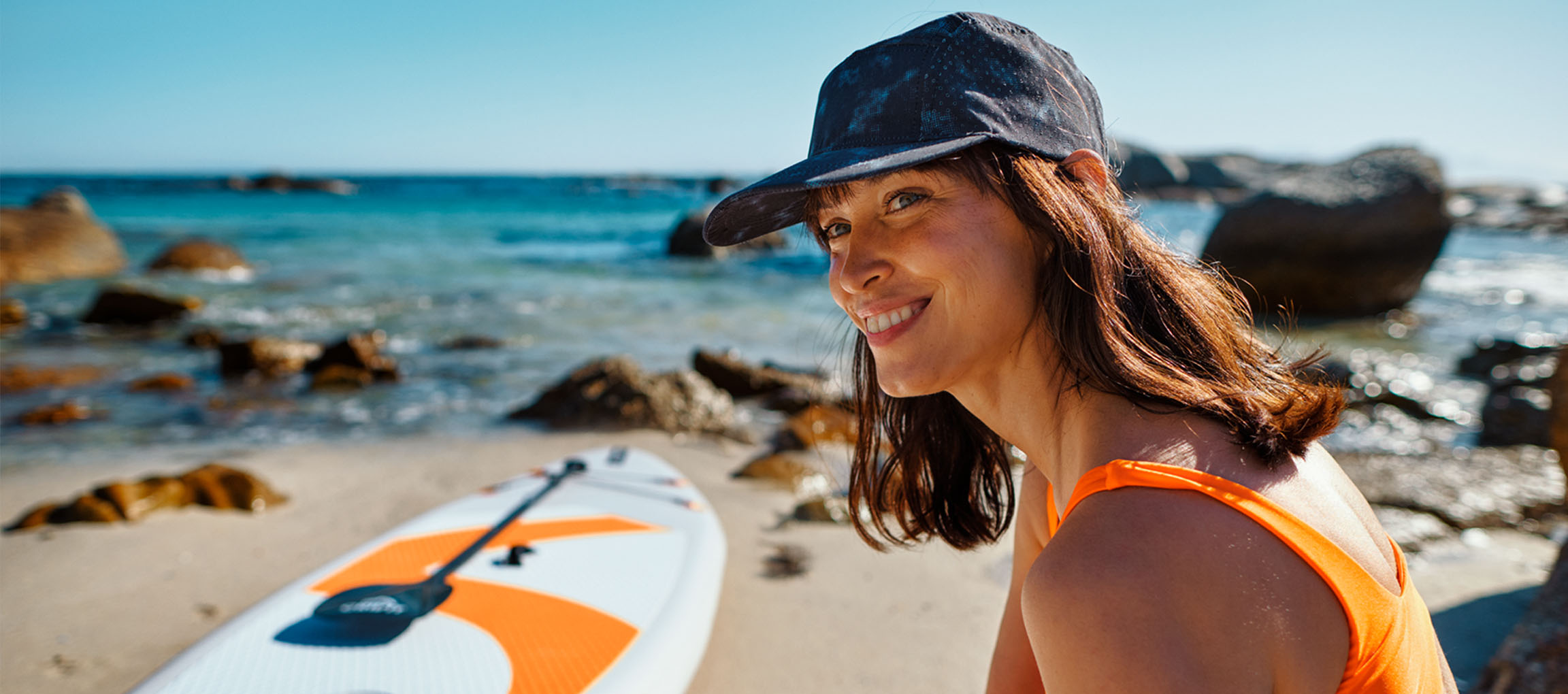 Smiling woman in orange swimsuit and baseball cap near a paddleboard on the beach.