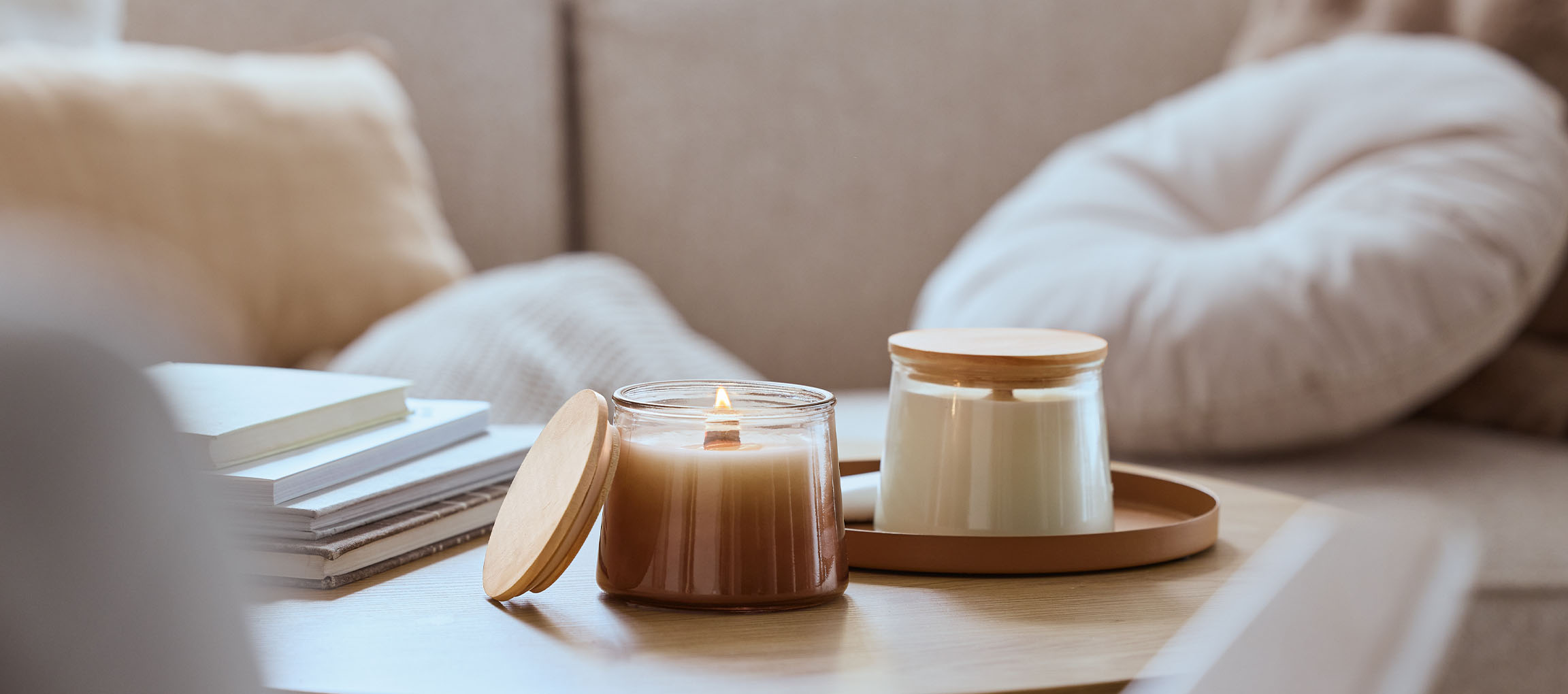 Two scented candles on a wooden table in a cozy living room with a sofa and cushions.