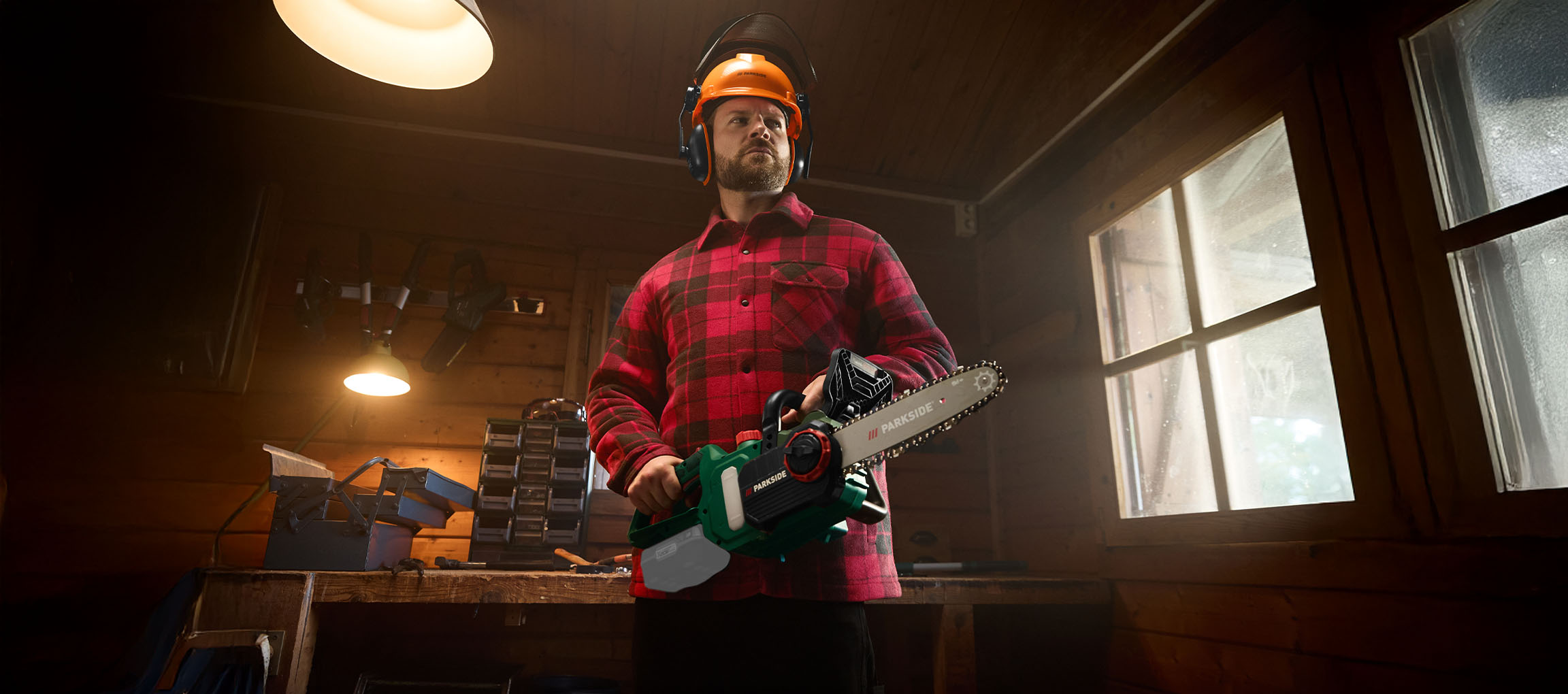 Man in a plaid shirt and safety gear holding a Parkside chainsaw in a workshop.