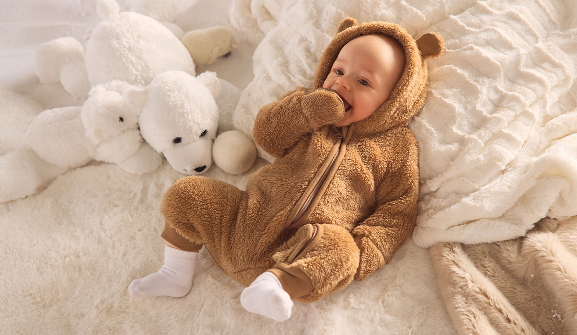 Baby in a brown bear onesie, lying on a white fluffy blanket with polar bear toys.