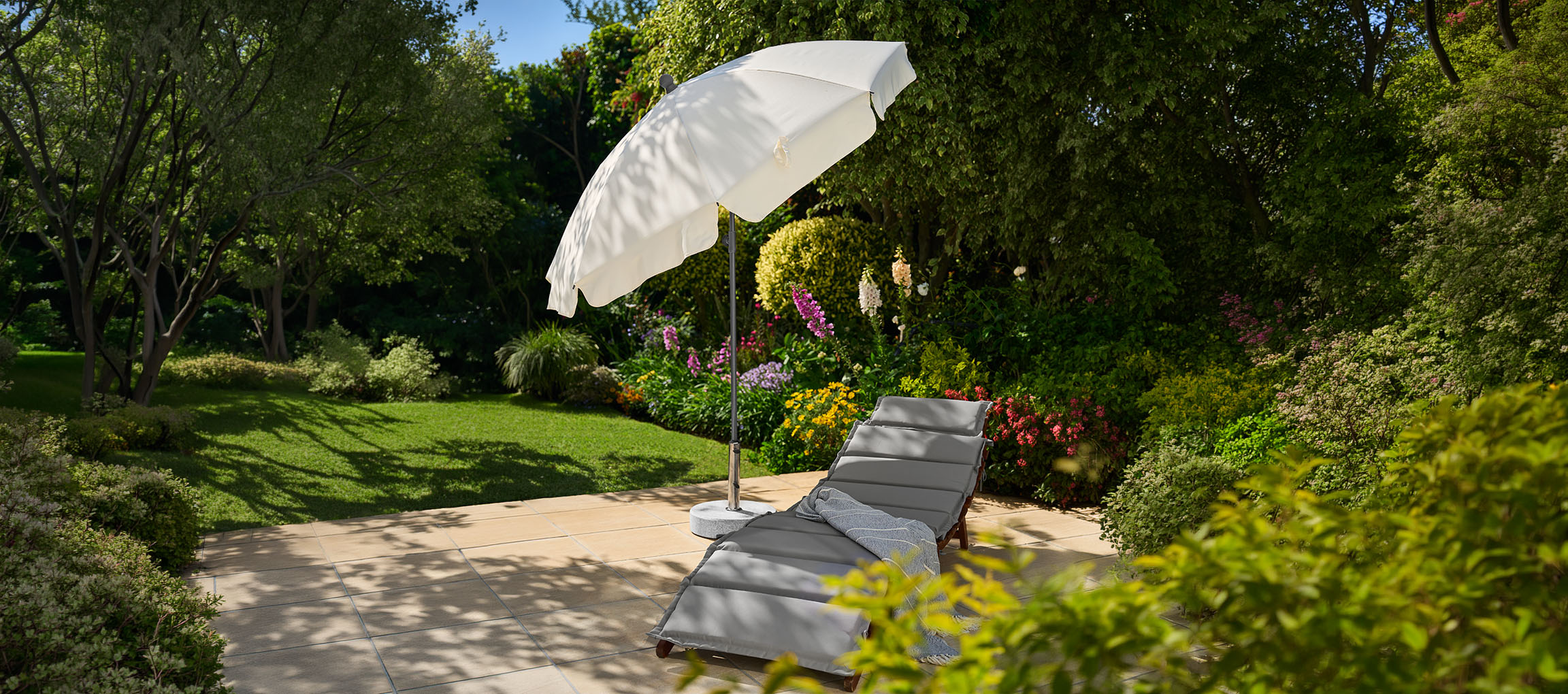 Garden sun lounger with umbrella on a patio, surrounded by greenery and flowers.
