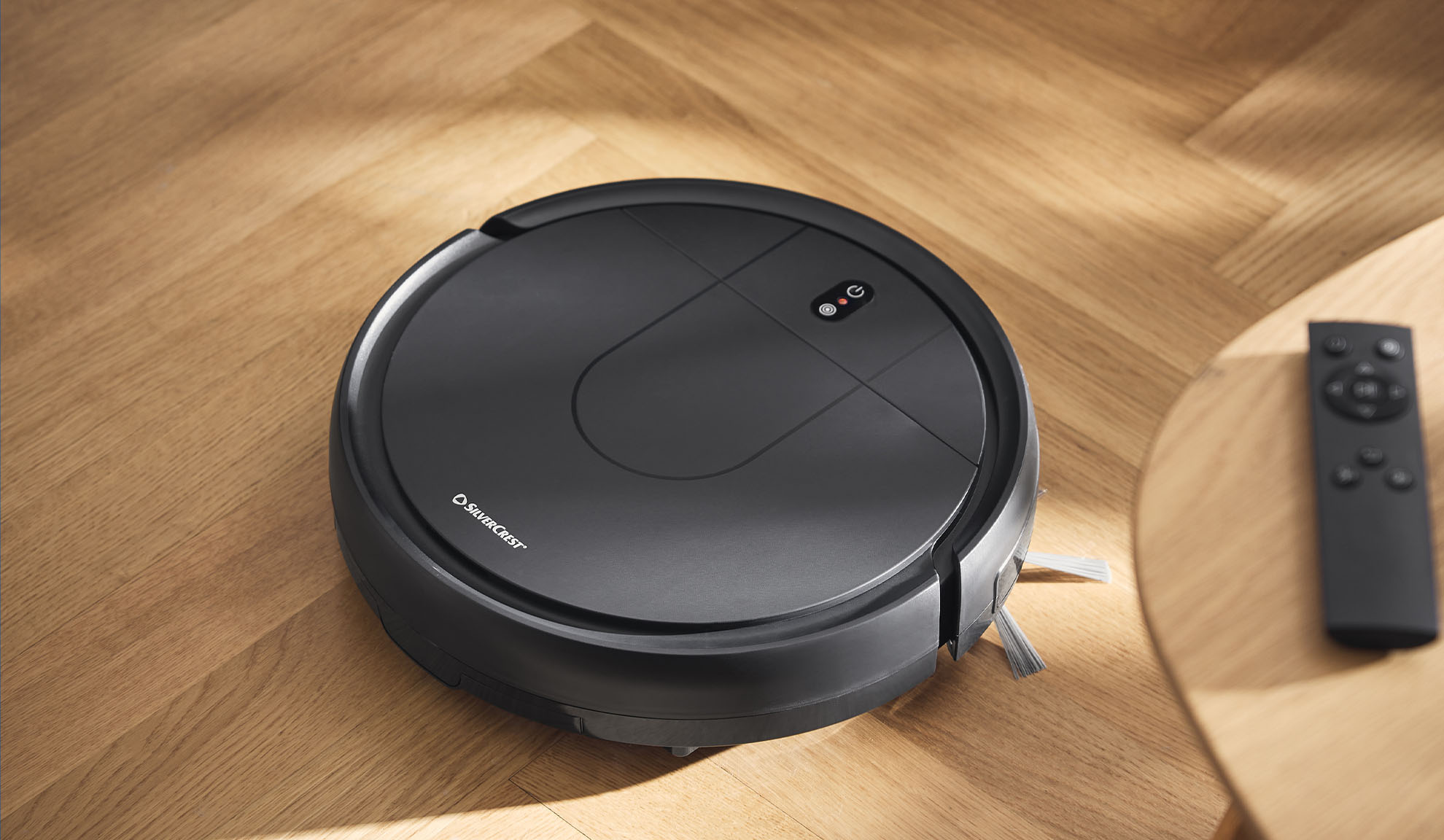 Robotic vacuum cleaner on a wooden floor with a remote control on a side table.