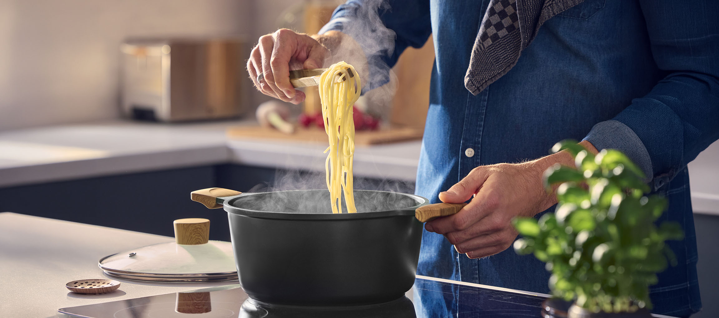 Man cooking pasta in a black pot with wooden handles, using wooden tongs.