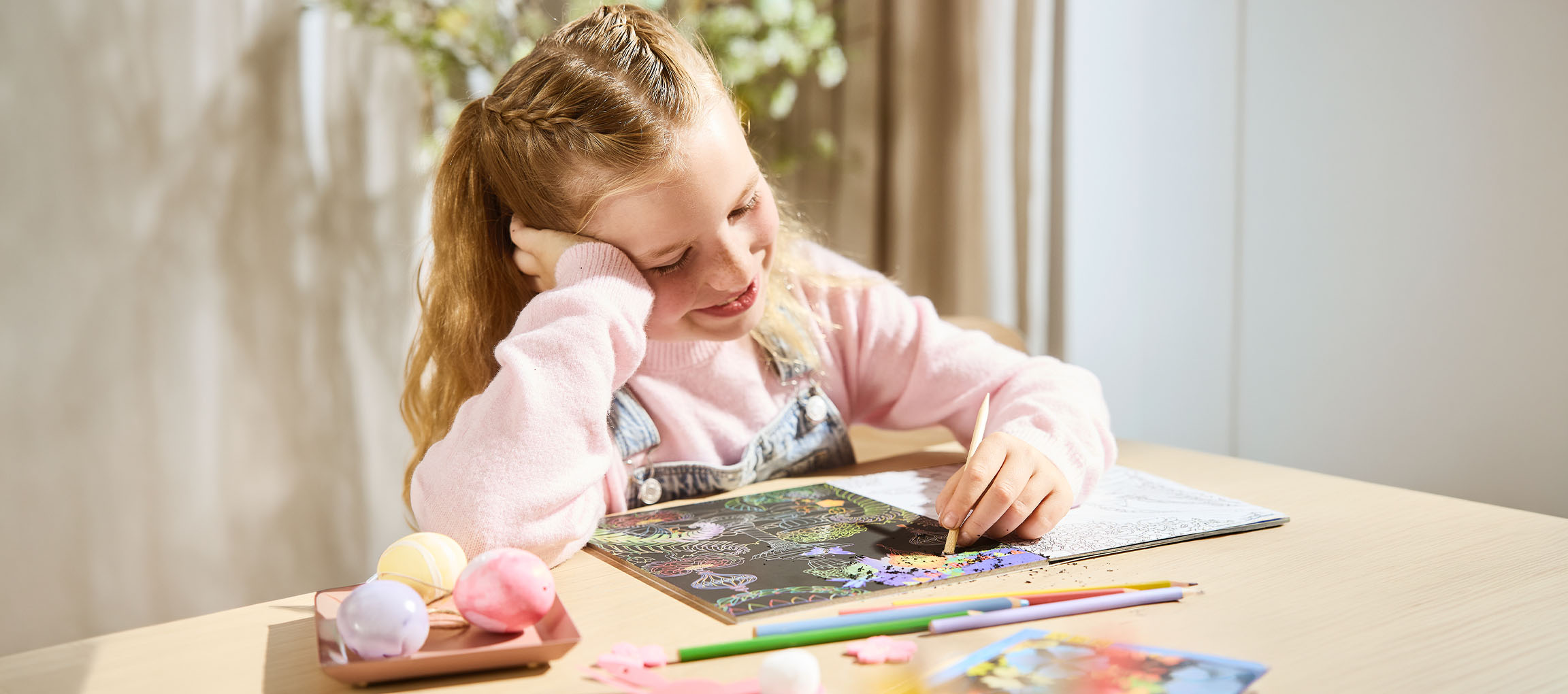 A girl scratching a colorful pattern onto a black scratch art paper, with colored pencils nearby.