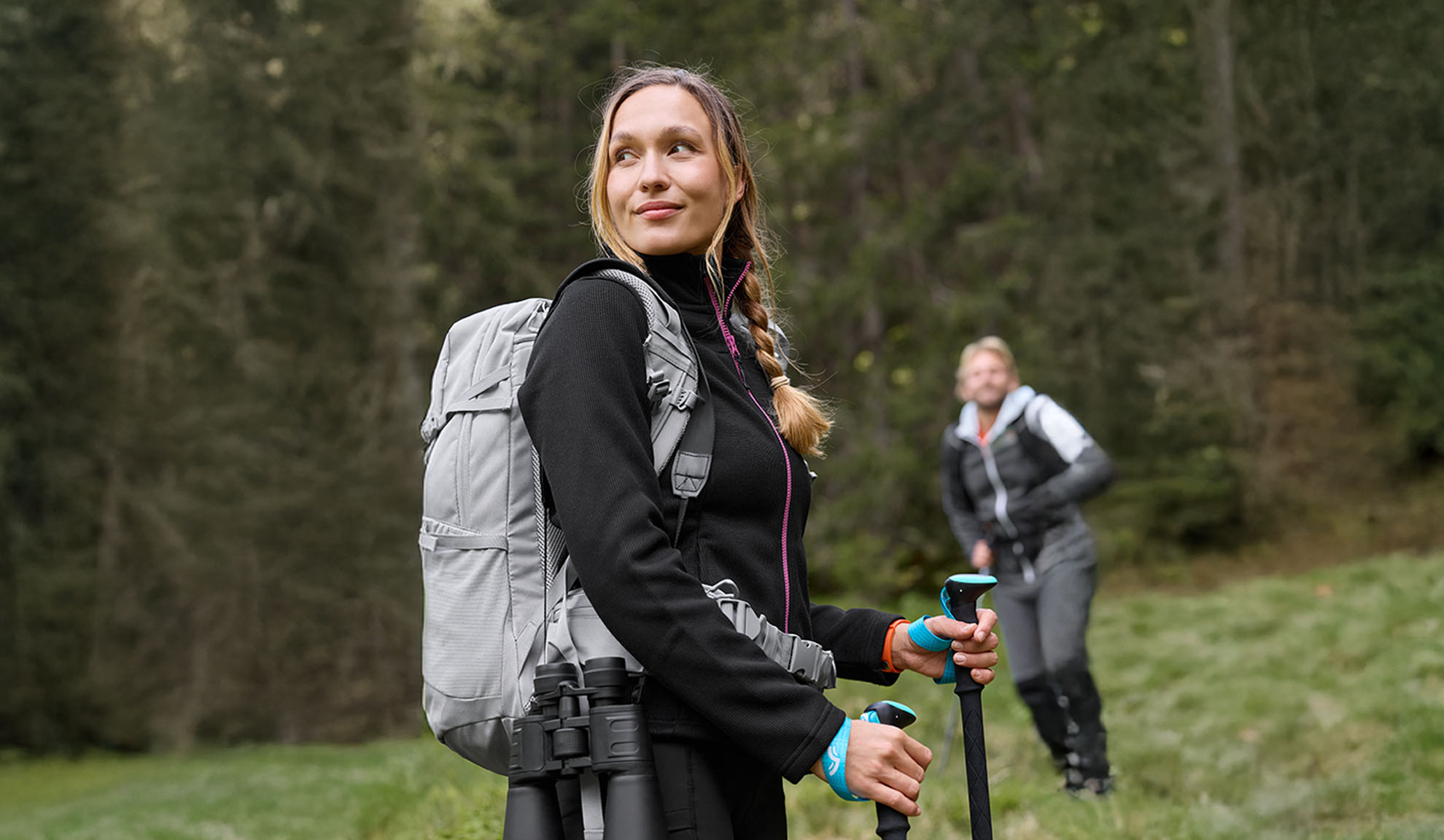 Woman with hiking backpack and trekking poles, man in background.