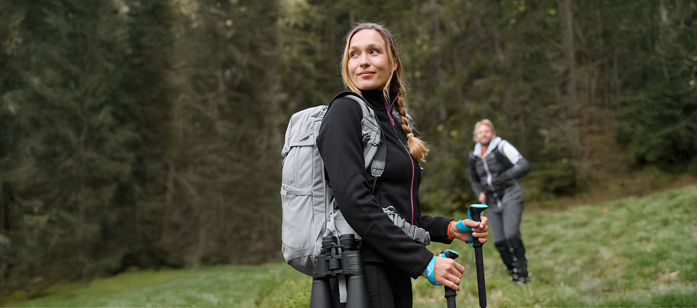 Woman with backpack and hiking poles, with a man in the background, in a forest.