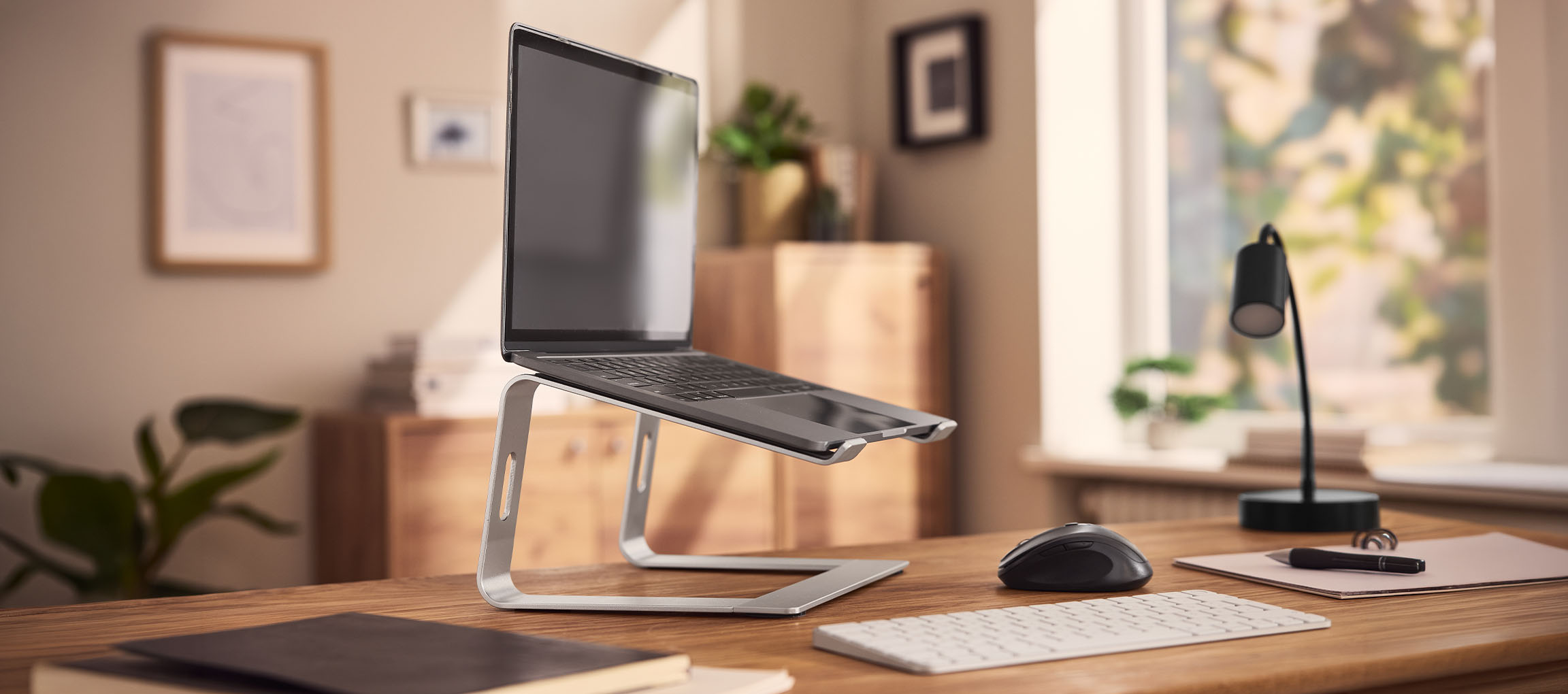 Laptop on a stand, mouse, and keyboard on a wooden desk in a bright room.