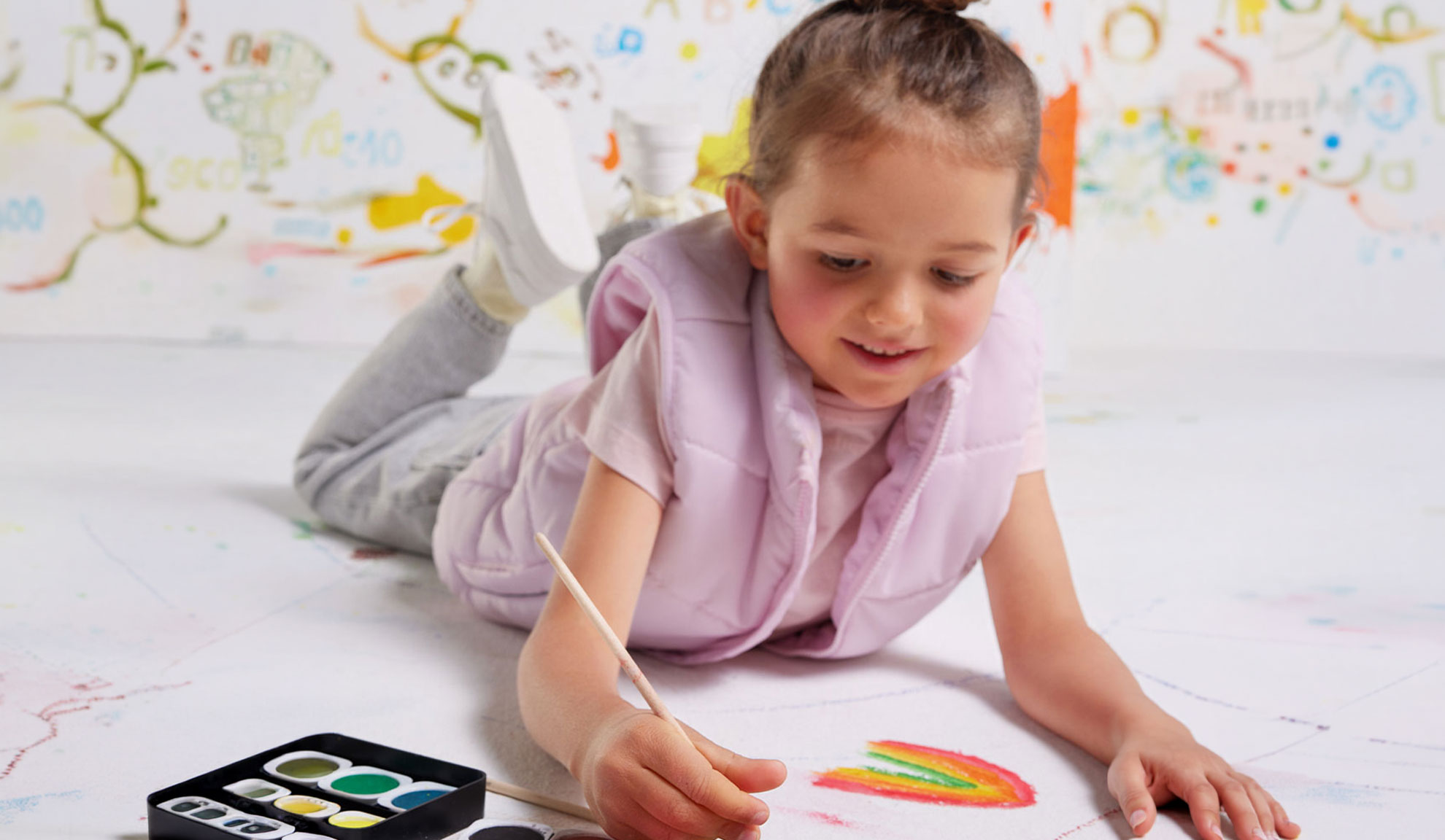A child painting a rainbow on paper with watercolors, wearing a pink vest.