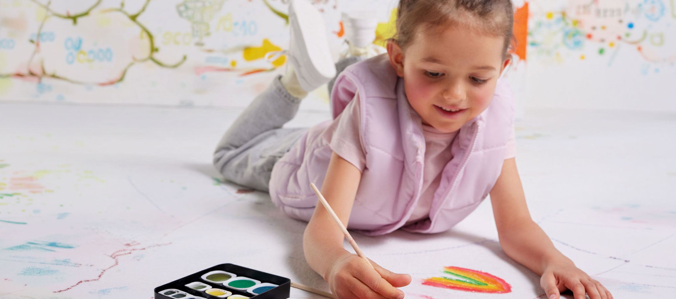 Girl painting with watercolors on the floor, wearing a pink vest.
