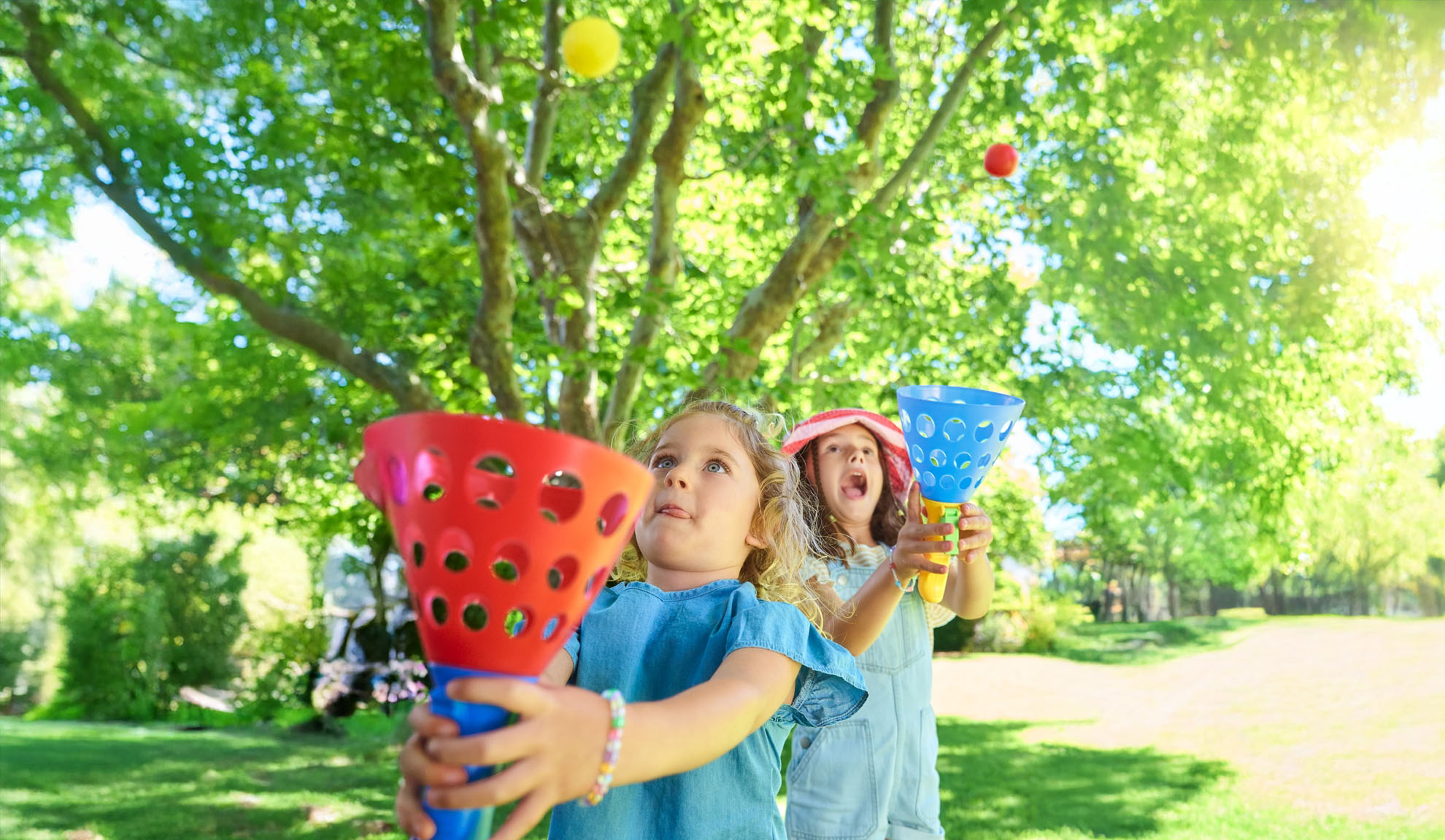 Two girls play a catching game outdoors with colorful plastic catchers and balls.