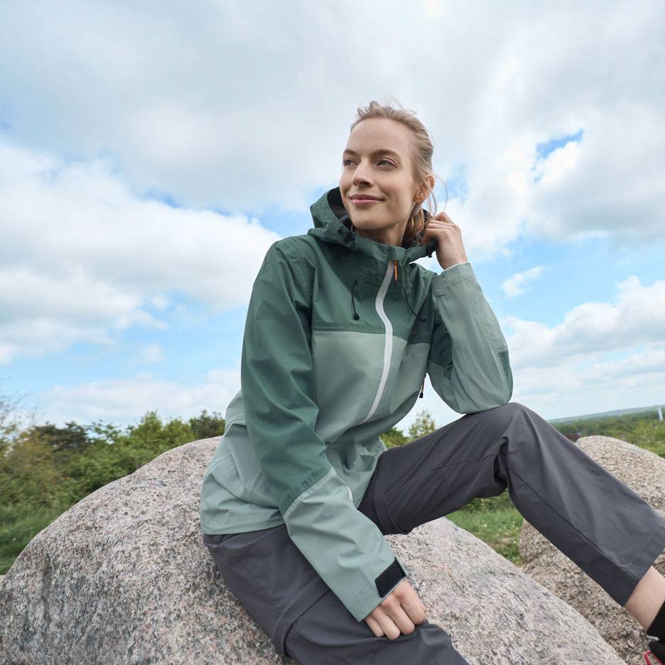 Woman in a green outdoor jacket and grey trousers sitting on a rock, looking into the distance.