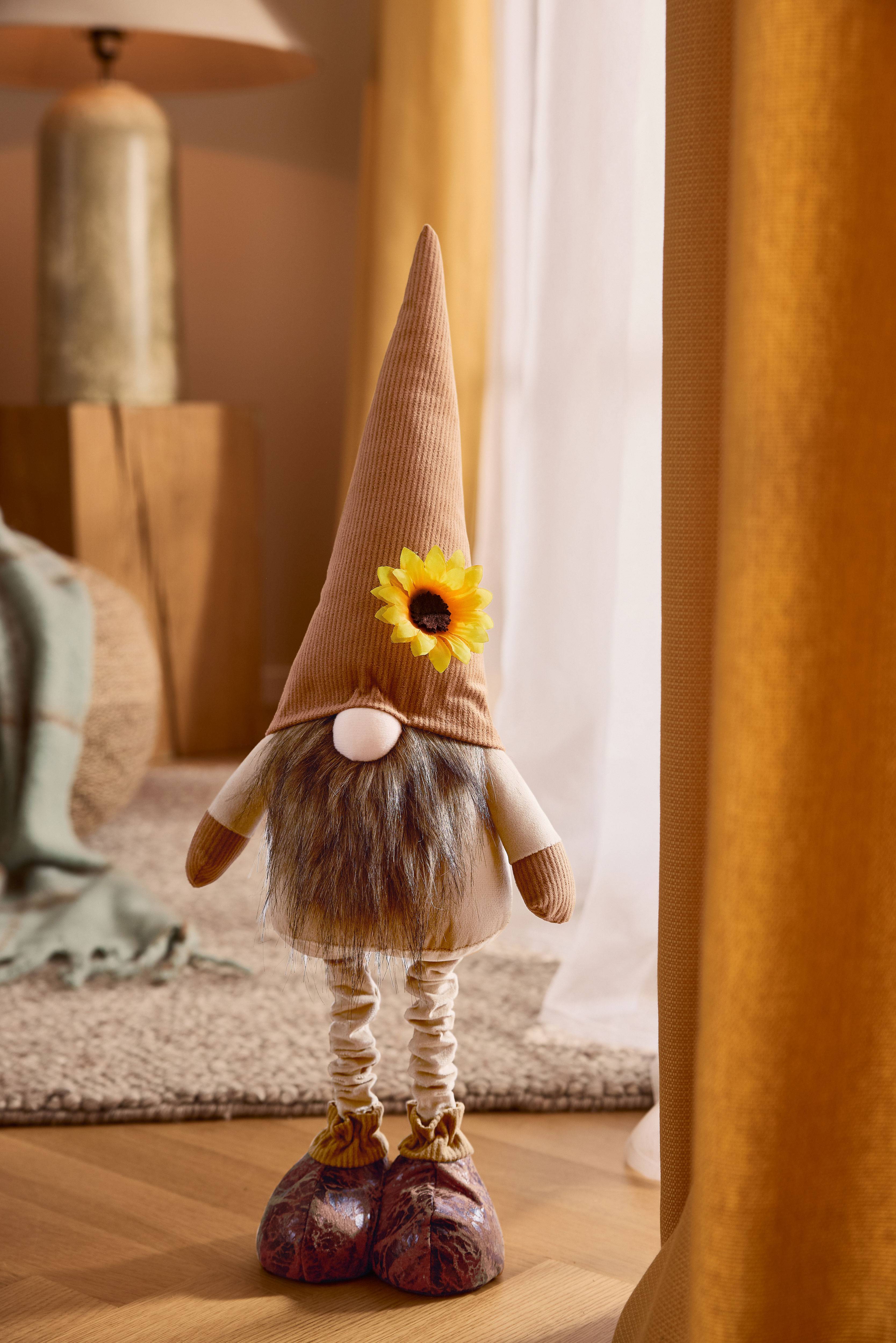 Plush gnome with a brown pointed hat, beard, and sunflower, in a cozy setting.