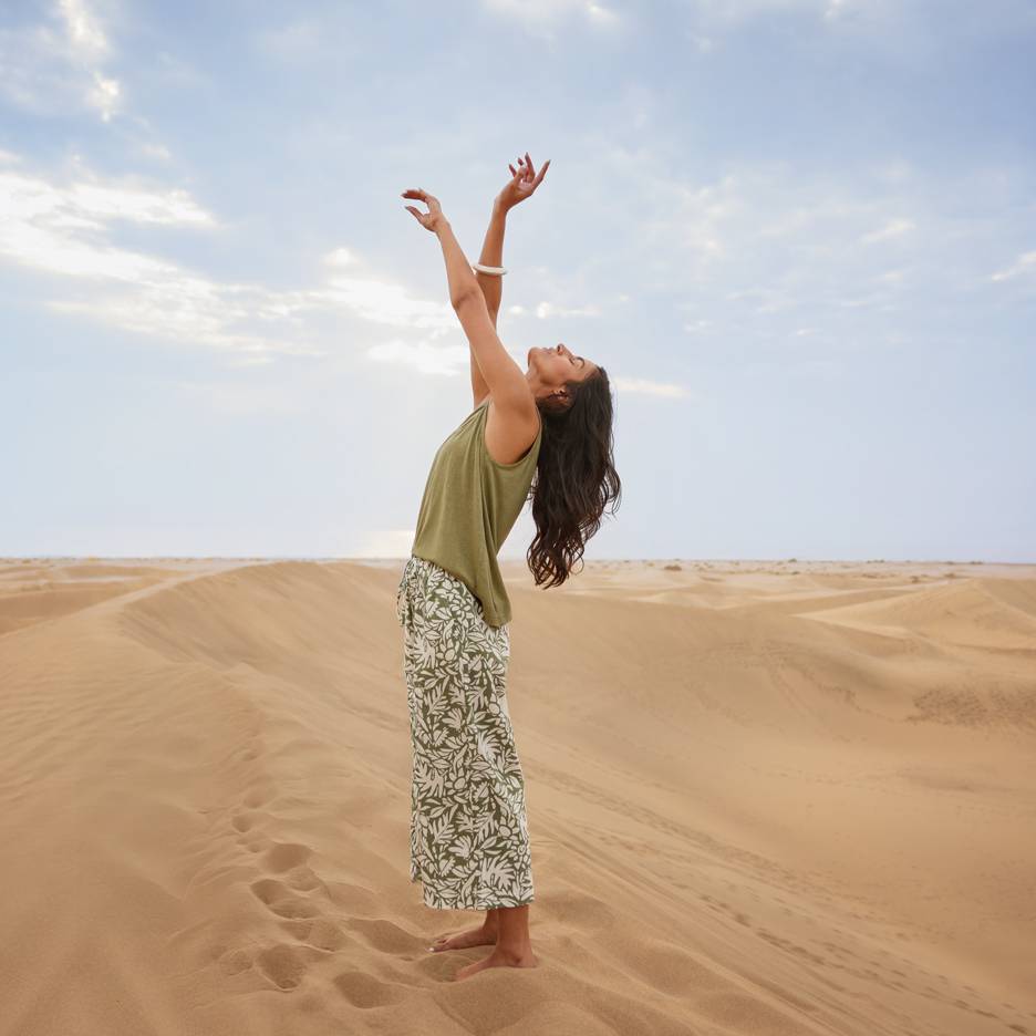 Woman in sand dunes wearing a green top and patterned trousers with raised arms.