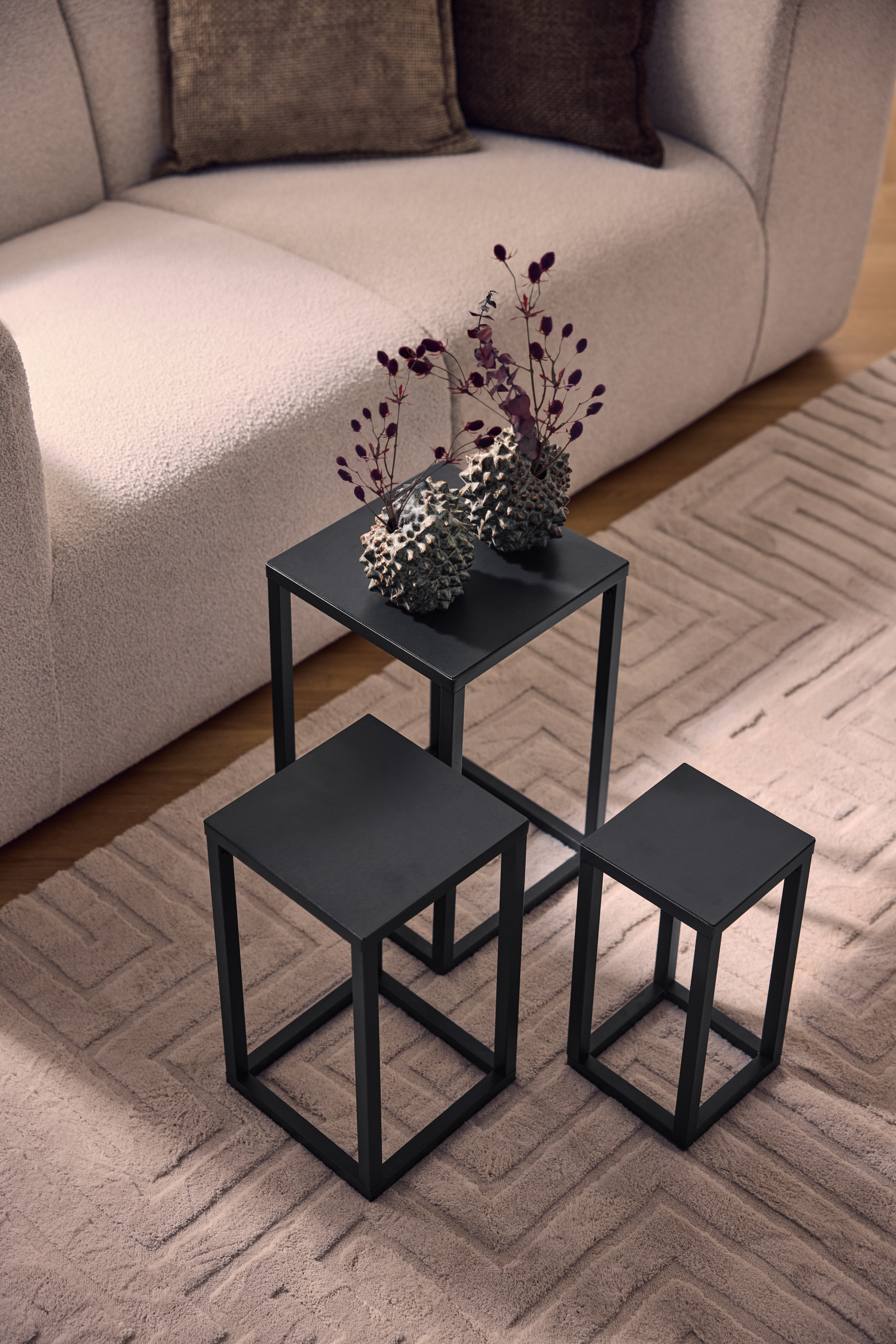 Three black metal coffee tables on a beige rug with a geometric pattern, next to a light sofa.